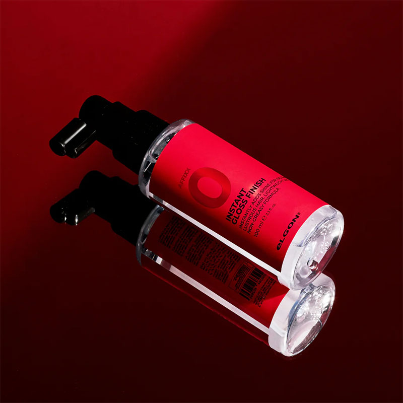 Flacone trasparente con etichetta rossa con scritte nere e tappo spray nero su  tavolo in vetro che riflette un telo rosso.